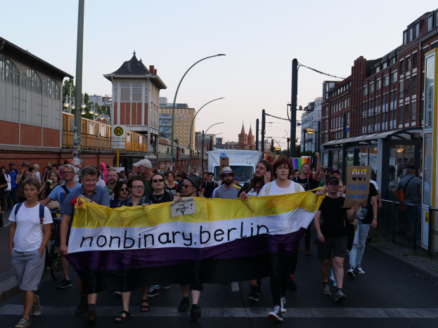 Gruppe von Menschen, die eine 'Nonbinary Berlin'-Fahne schwenken, auf einer Berliner Straße mit Laternen, Gebäuden, Bushaltestelle, Bäumen und einem klaren blauen Himmel im Hintergrund.