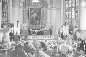 Schwarzes und weißes Foto einer Trauerfeier in einer Kirche mit sitzenden und stehenden Menschen, Blumenbouquets, Kerzen, einem Sarg im Vordergrund, gerahmten Fotos und Skulpturen an der Wand und Fenstern auf beiden Seiten.