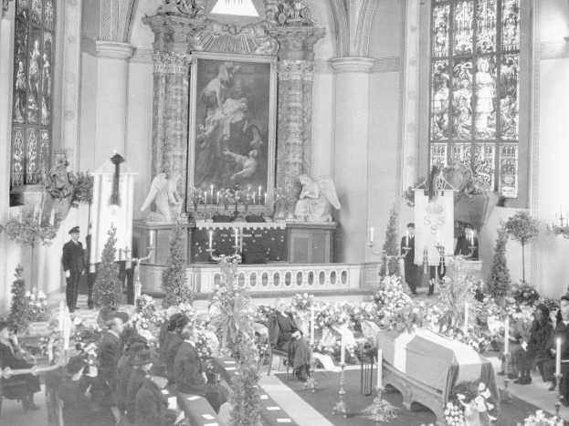 Schwarzes und weißes Foto einer Trauerfeier in einer Kirche mit sitzenden und stehenden Menschen, Blumenbouquets, Kerzen, einem Sarg im Vordergrund, gerahmten Fotos und Skulpturen an der Wand und Fenstern auf beiden Seiten.