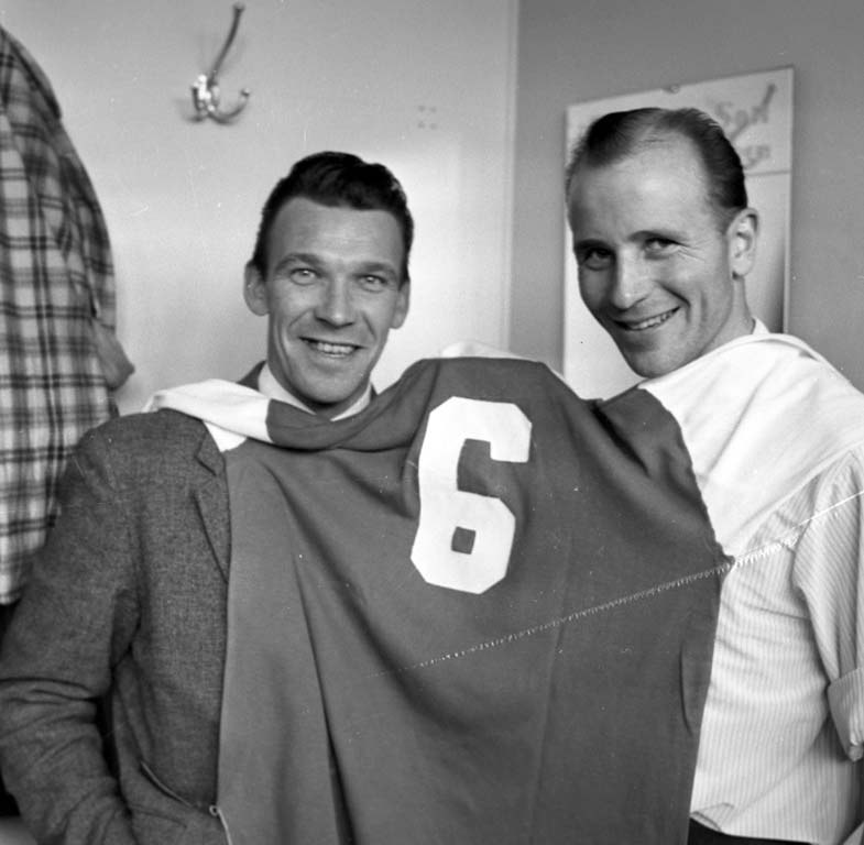 Zwei Männer in Schwarz und Weiß, die strahlend eine Fußballtrikot hochhalten, mit einer Wandtafel und einem Haken im Hintergrund.
