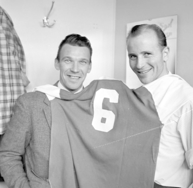Zwei Männer in Schwarz und Weiß, die strahlend eine Fußballtrikot hochhalten, mit einer Wandtafel und einem Haken im Hintergrund.