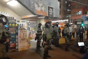 Polizeibeamte in Schutzausrüstung stehen vor einer Menge von Helikopter-Protestlern mit Waffen, mit Plakaten an Wänden, Deckenleuchten, Gebäuden und einer Verkehrsampel mit Text im Hintergrund, in Hong Kong.