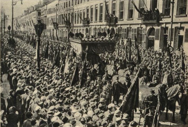 Eine gro├če Menge Menschen marschiert auf einer Stra├če vorbei, mit Fahnen tragenden Menschen, Geb├Ąuden, Laternenmasten und einem klaren Himmel im Hintergrund und dem Text "1918 - der Umzug der Truppen" unten.