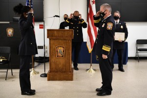 Eine Gruppe uniformierter Personen in Masken steht vor einem Podium mit einem Mikrofon, umgeben von Flaggen, Stühlen, Tischen und einer Wand, wobei eine Person ein Zertifikat hält, während es scheint, dass eine Amtseinführung für einen neuen Feuerwehrchef stattfindet.