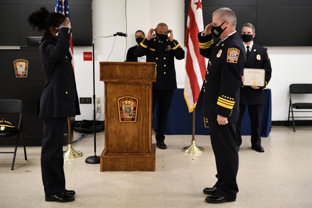 Eine Gruppe uniformierter Personen in Masken steht vor einem Podium mit einem Mikrofon, umgeben von Flaggen, Stühlen, Tischen und einer Wand, wobei eine Person ein Zertifikat hält, während es scheint, dass eine Amtseinführung für einen neuen Feuerwehrchef stattfindet.