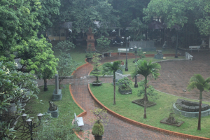 Ein grüner Park mit Bäumen, Pflanzen, Gras, Laternen, Bänken, Skulpturen und einem Gebäude im Hintergrund.