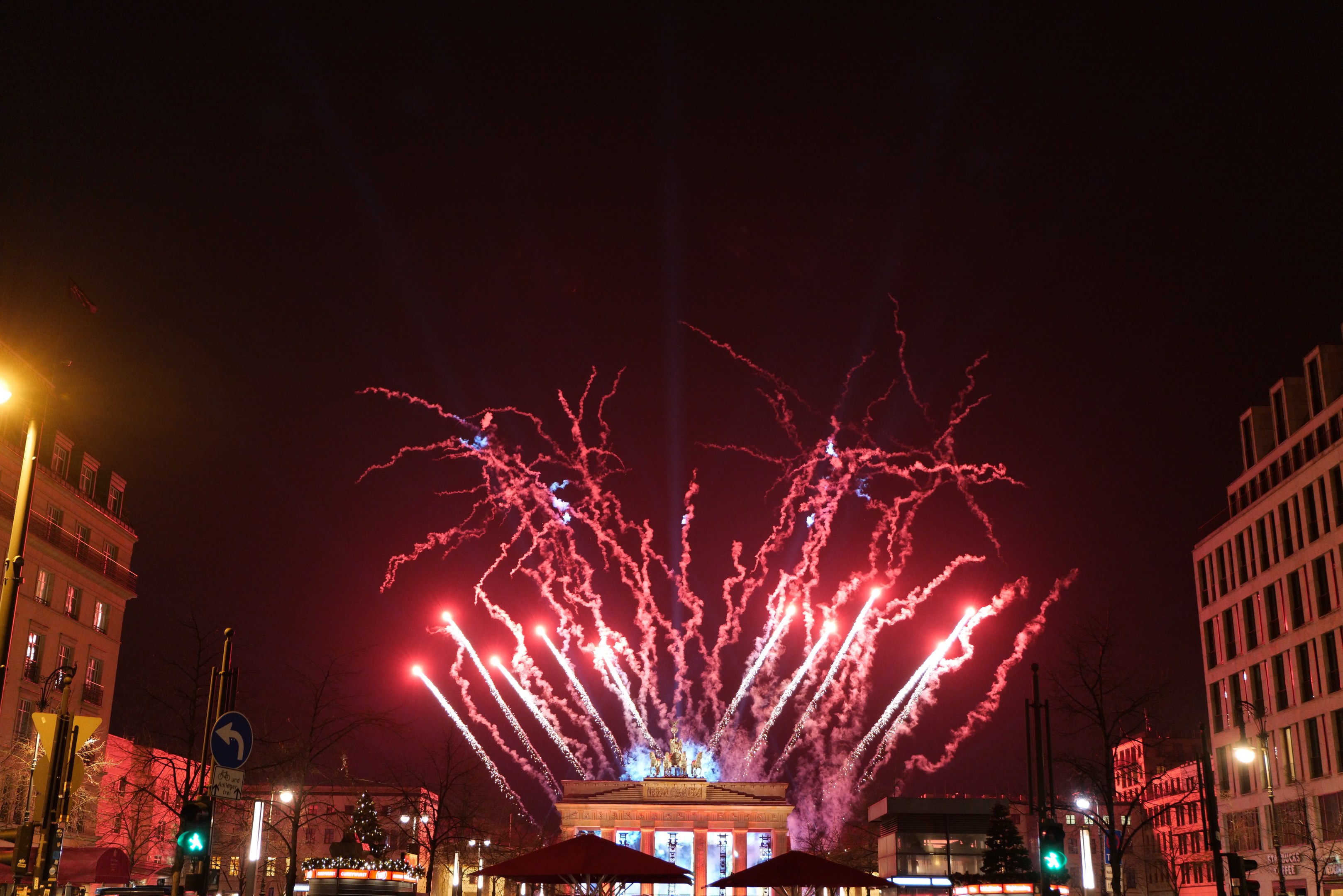 Eine belebte Stadtstraße am Silvesterabend in Berlin, voller Menschen, Fahrzeuge, Bäume, Pfosten, Lichter, Schilder, Verkehrszeichen und Gebäude, beleuchtet von Gebäudelichtern und Feuerwerk am Himmel.