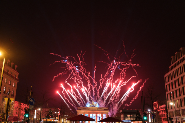 Eine belebte Stadtstraße am Silvesterabend in Berlin, voller Menschen, Fahrzeuge, Bäume, Pfosten, Lichter, Schilder, Verkehrszeichen und Gebäude, beleuchtet von Gebäudelichtern und Feuerwerk am Himmel.