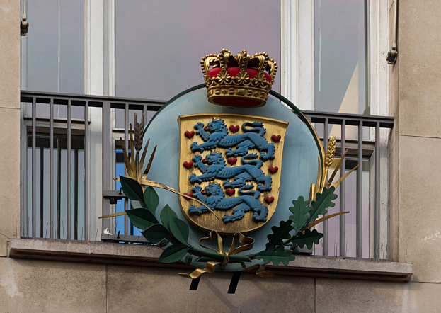 Wappen der britischen Königsfamilie an einer Gebäudefassade mit einer Krone- und Blätter-Logo, Fenstern und Metallgittern im Hintergrund.