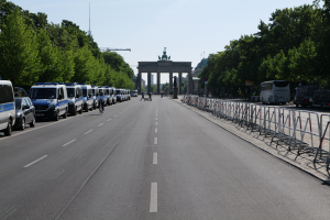 Eine Reihe von Polizeiautos auf einer Straße vor dem Brandenburger Tor in Berlin, Deutschland, mit Menschen auf Fahrrädern und Stehenden, Barrieren, Bäumen und einem Bogen mit Statuen im Hintergrund.