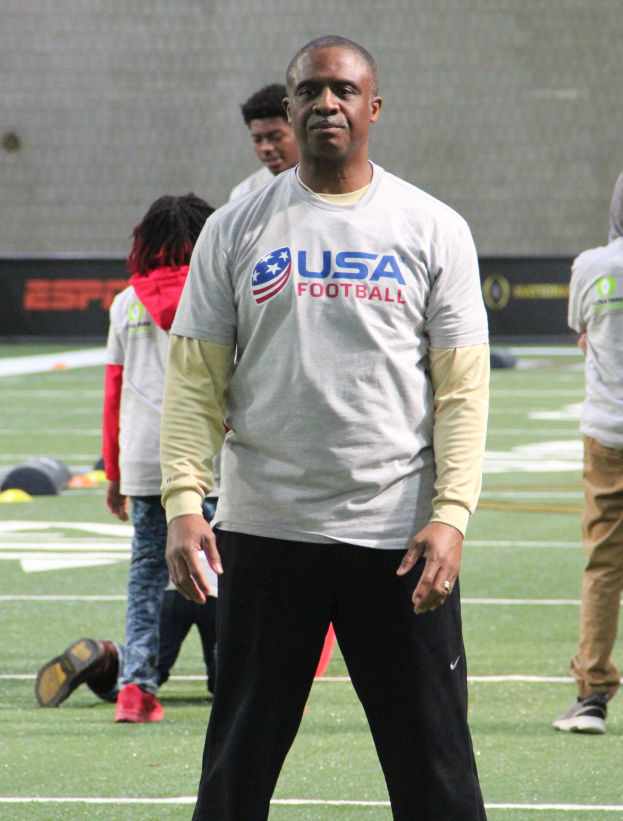 Ein Fußballspieler steht auf einem Feld mit einer Gruppe von Menschen und einer Wand im Hintergrund.