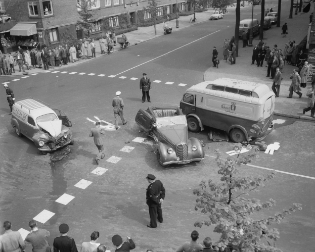Schwarz-weißes Bild eines Autounfalls an einer Straßenkreuzung mit mehreren Fahrzeugen, darunter ein Lieferwagen, und einer Gruppe von Zuschauern; Gebäude, Bäume, Masten und Texttafeln sind im Hintergrund zu sehen.