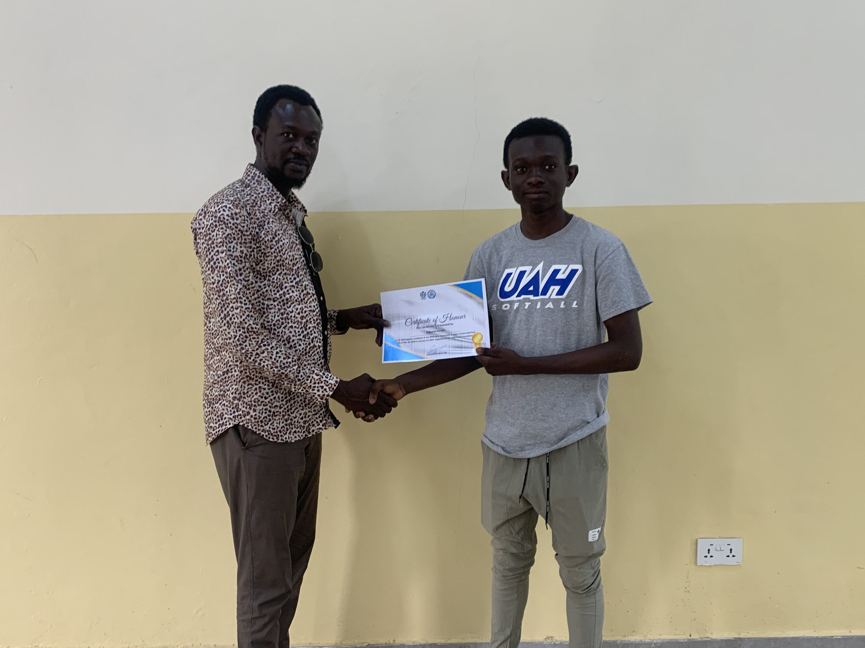 Zwei Männer, die vor einer gelben Wand Hände schütteln, wobei der Mann links ein Zertifikat hält, das er von der Universität Ghana erhalten hat.