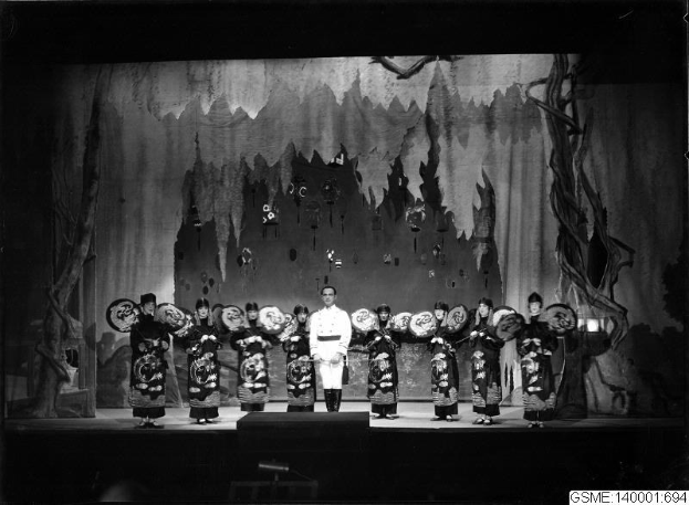 Schwarzes und weißes Foto von Darstellern in traditionellen japanischen Kimonos auf einer Bühne, die Gegenstände halten, mit einer bemalten Kulisse und Text, der unten 'Kimono-Show in Tokyo, Japan' liest.