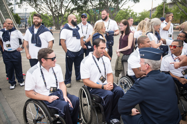 Eine Gruppe von Menschen im Rollstuhl unterhält sich vor einer Kulisse aus Bäumen, Pfählen, Gebäuden und einem klaren blauen Himmel, wobei einige Mützen und Brillen tragen, was auf eine Teilnahme an den Paralympischen Spielen hindeutet.