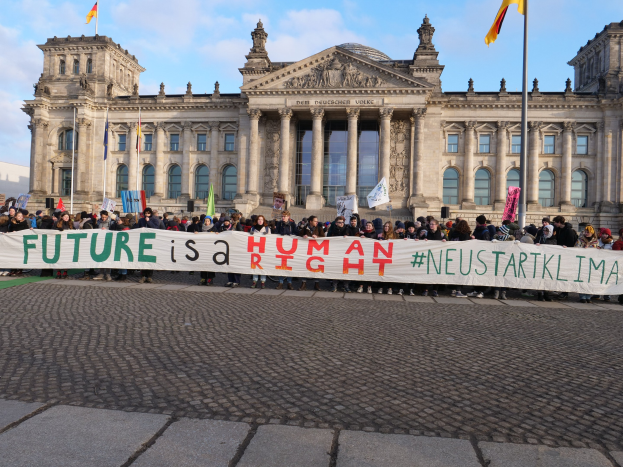 Eine Gruppe von Menschen mit einem Transparent 'Zukunft ist ein Mensch' vor dem Reichstaggebäude in Berlin, Deutschland, mit sichtbaren architektonischen Details und Flaggen im Hintergrund bei bewölktem Himmel.