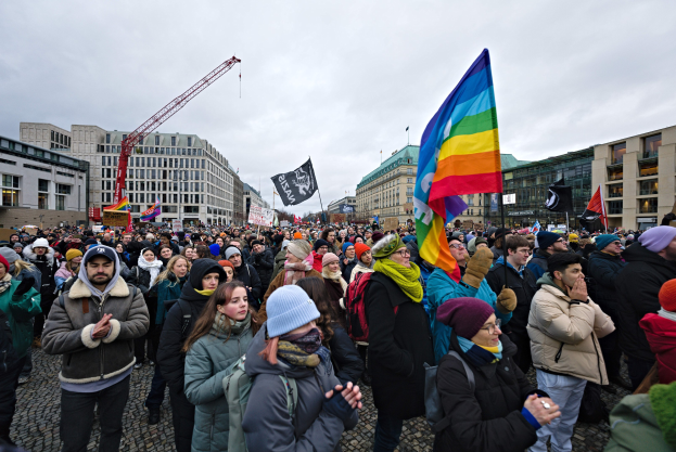 Eine große Gruppe von Menschen bei einer LGBTQ+-Rechtsmarsch in Berlin, die Fahnen und Plakate schwenken, mit Gebäuden, einem Kran und einem bewölkten Himmel im Hintergrund.