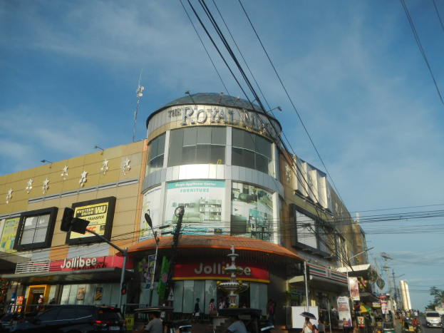 Eine belebte Stadtstraße mit Fahrzeugen auf der Straße, Fußgängern auf dem Gehweg und dem Royal Mall Einkaufszentrum im Hintergrund, umgeben von Bäumen und Strommasten.