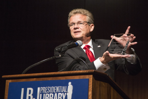 Ein Mann in Anzug und Krawatte steht an einem Rednerpult und hält eine Auszeichnung in der Hand, mit einem Mikrofon und einer Tafel daran, trägt eine Brille vor einem dunklen Hintergrund bei einer Preisverleihung in einer Bibliothek.