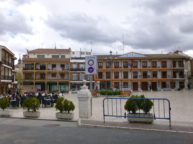 Ein belebter Stadtplatz mit Menschen, die sitzen und stehen, Topfpflanzen, eine Metallabsperrung, ein Schild an einem Pfahl, Straßenlaternen mit Fahnen, umliegende Gebäude und ein bewölkter Himmel.