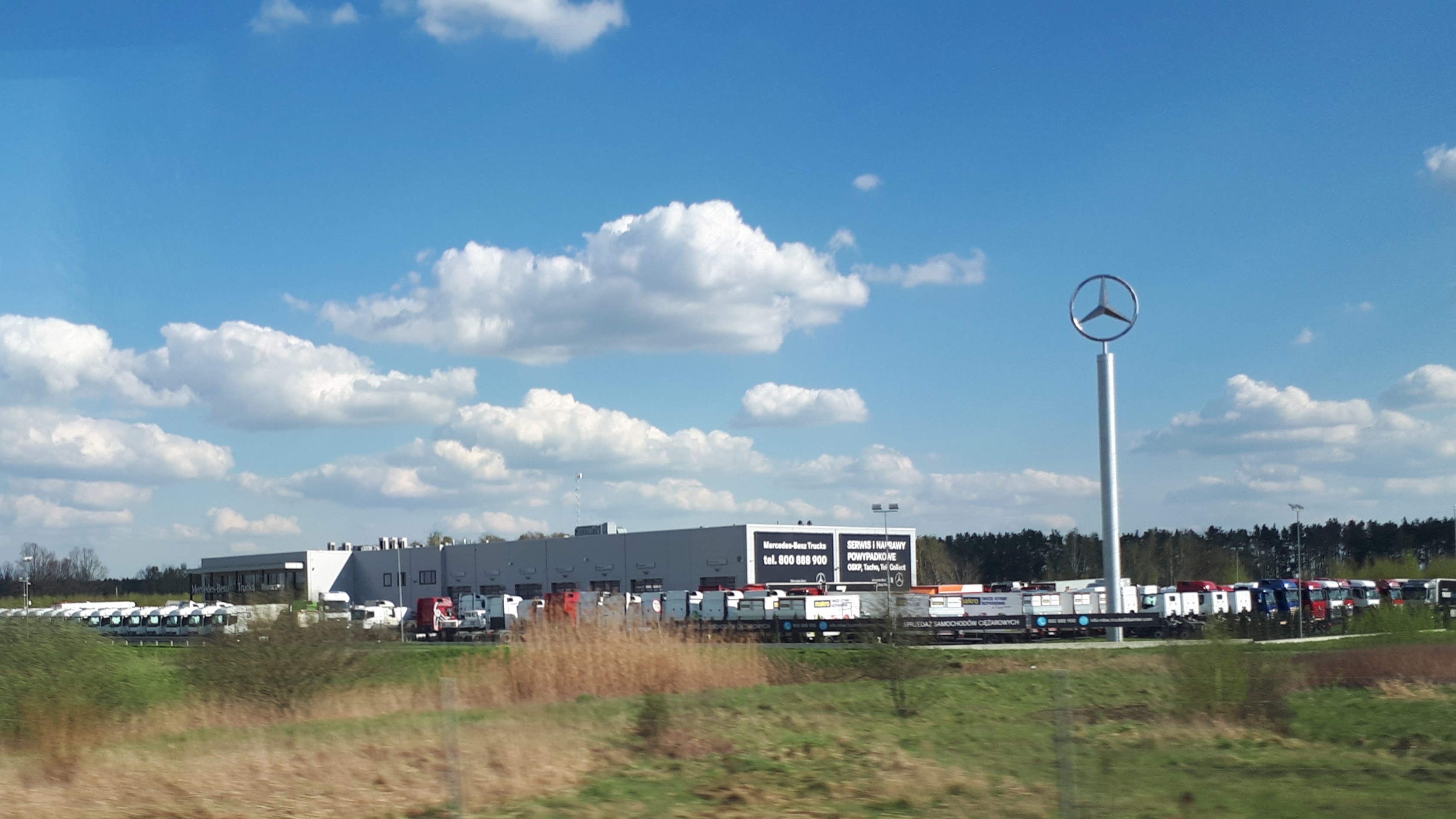 Mercedes-Benz-Fabrik in Deutschland mit Fahrzeugen auf der Straße, Grünfläche, Gebäuden und einem Logo-Mast unter einem bewölkten Himmel.
