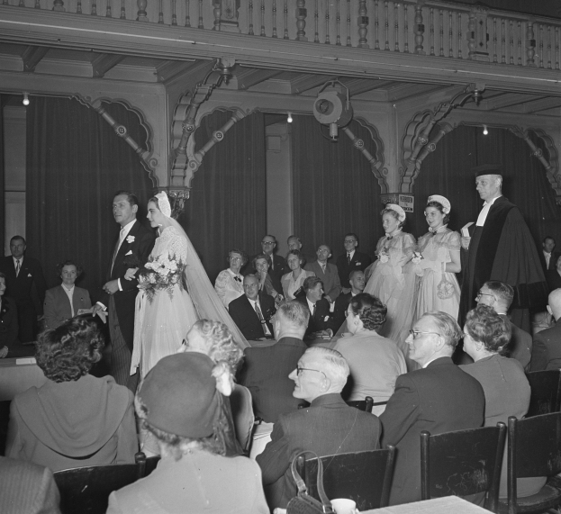 Ein Schwarz-Weiß-Foto einer Hochzeitszeremonie in einem großen Raum mit vielen sitzenden und stehenden Gästen, Sträußen, Tassen auf Tischen und Vorhängen mit Deckenlampen im Hintergrund.