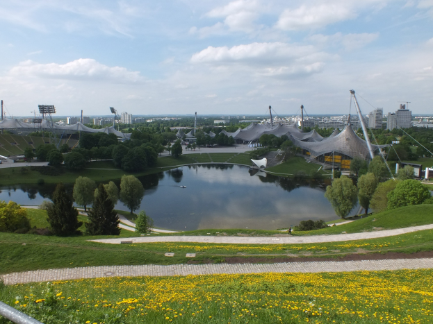 Luftaufnahme des Olympiastadions von einem Hügel aus, mit einem Teich im Vordergrund, der von Grünflächen und gelben Blumen umgeben ist, und Gebäuden im Hintergrund unter einem klaren blauen Himmel.