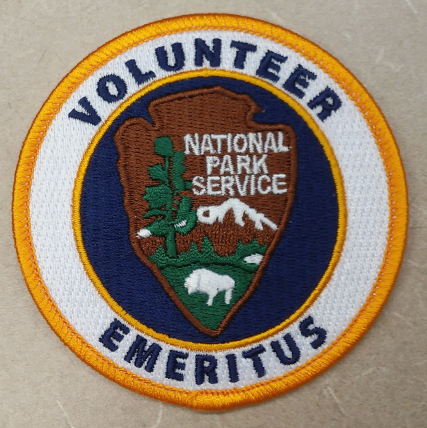 Runder Aufnäher mit blauem Rand und weißem Hintergrund, darauf in schwarzer Schrift "Volunteer National Park Service Emeritus" in der Mitte eines gelben Kreises.