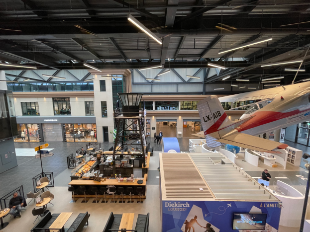 Ein geräumiger Raum mit zahlreichen Tischen, Stühlen, Sofas und verschiedenen Gegenständen, einschließlich eines Flugzeugs auf der rechten Seite, mit einigen anwesenden Personen und Glasfenstern und -wänden im Hintergrund.