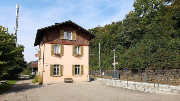 Ferienhaus zum Verkauf in Baden-Württemberg, Deutschland, mit einem Gebäude mit zahlreichen Fenstern, einem vorderen Zaun, Straßenmöbeln, Grünfläche und einem bewölkten Himmel.