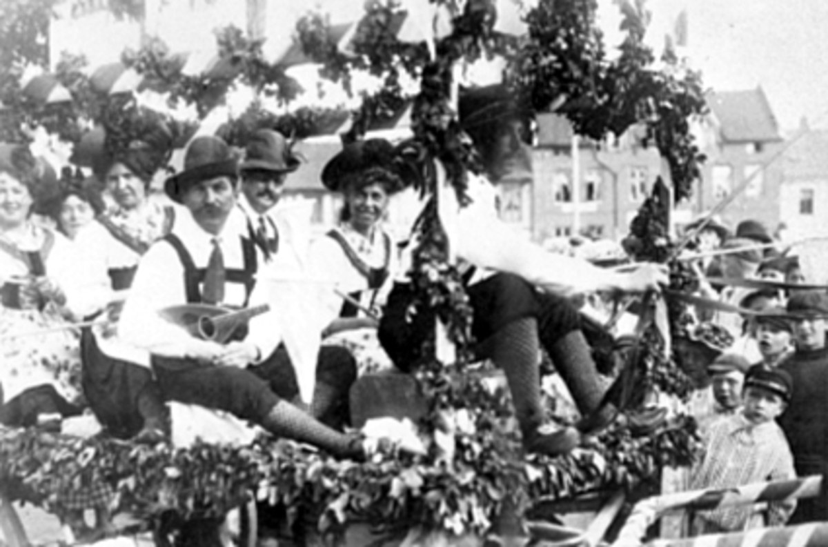 Schwarzes und weißes Bild eines Paradewagens mit Menschen in traditioneller bayrischer Tracht, einige spielen Instrumente, mit Blattdekoration vor einem Hintergrund aus Gebäuden und einem klaren Himmel.