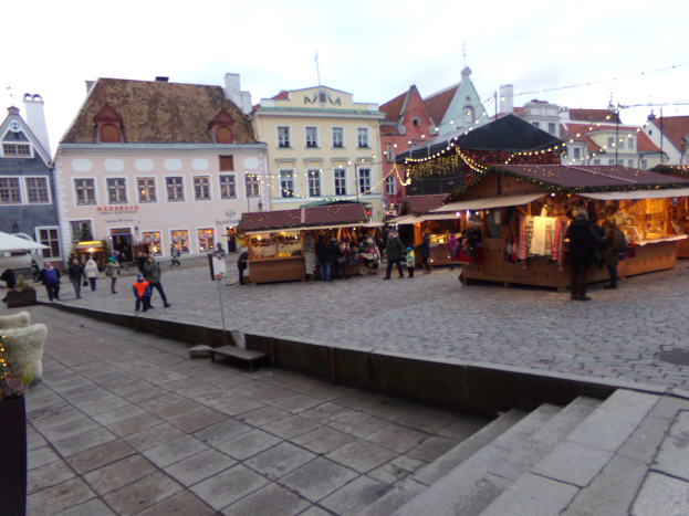 Ein geschäftiger Weihnachtsmarkt in Tallinn, Estland mit Menschen um geschmückte Stände, festliche Lichter, bewölkten Himmel, Gebäuden im Hintergrund, Treppen nach oben und Topfpflanzen.