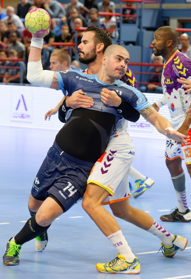 Mannergruppe spielt Handball in einem Stadion, einer hält den Ball, mit Zuschauern im Hintergrund.