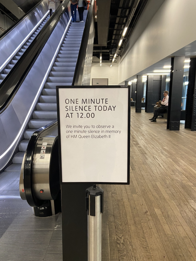 Eine Rolltreppe im Flughafen mit einem Schild, auf dem "Eine Minute Stille heute" steht, einige Menschen darauf und Lampen an der Decke im Hintergrund.