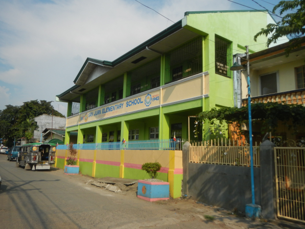 Schulgebäude mit grüner und gelber Fassade, Straße mit Fahrzeugen, Topfpflanzen, Zaun, Tor, Straßenlaterne, Bäume, Drähte und bewölkter Himmel.
