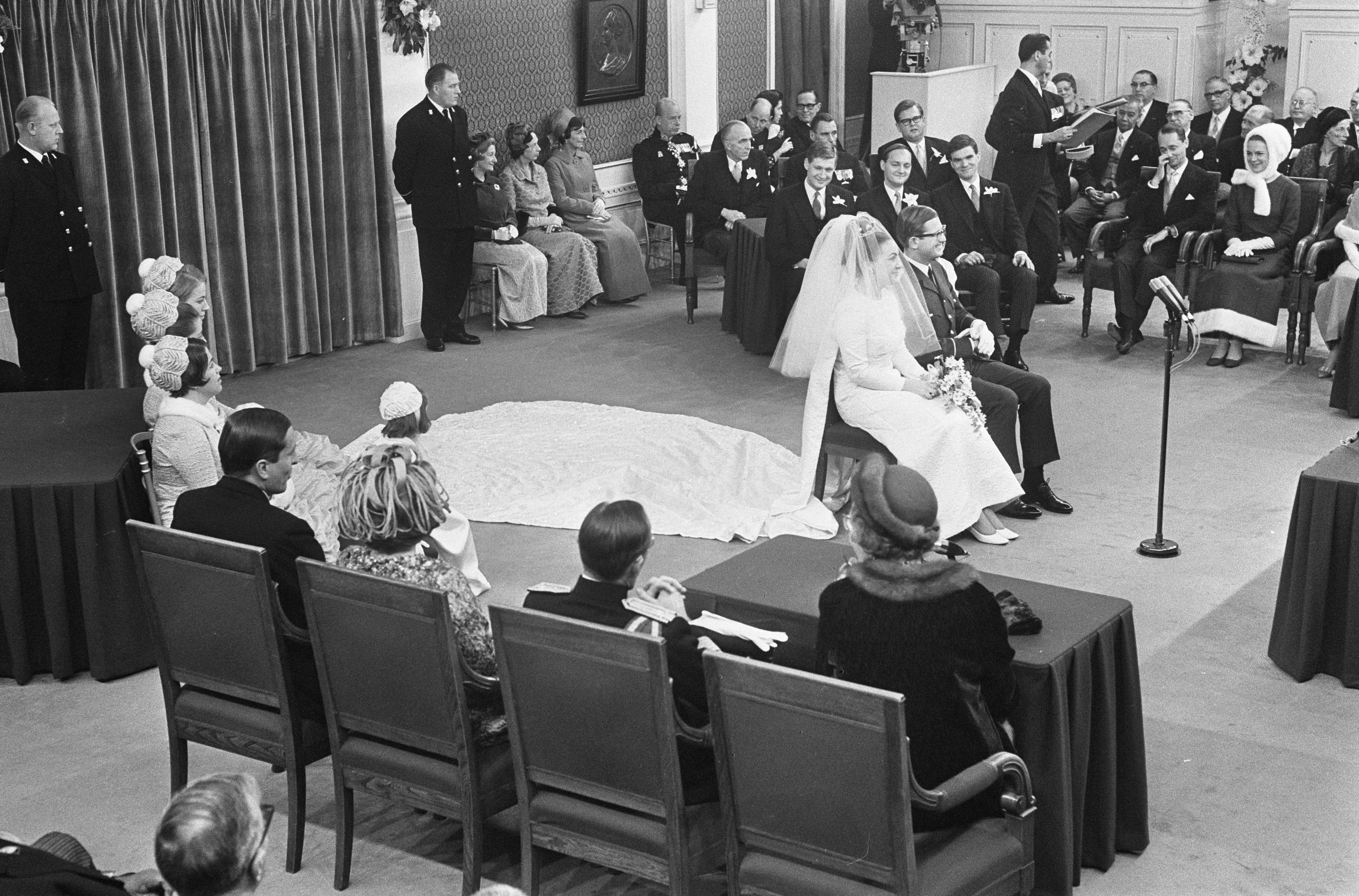 Schwarzes und weißes Foto einer Hochzeitszeremonie mit Menschen, die sitzen und stehen, einem Mann mit Mikrofon, einer Frau mit Blumenstrauß, einem Podium mit Mikrofon, Blumengestecken, einem Vorhang, einem gerahmten Foto an der Wand und einer Tür im Hintergrund.