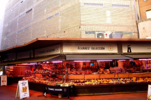 Ein belebter Markt mit verschiedenen Lebensmitteln, beleuchtet von Deckenlampen, umgeben von Texttafeln, vor Gebäuden unter einem klaren blauen Himmel.