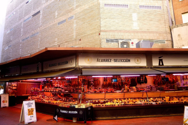 Ein belebter Markt mit verschiedenen Lebensmitteln, beleuchtet von Deckenlampen, umgeben von Texttafeln, vor Gebäuden unter einem klaren blauen Himmel.