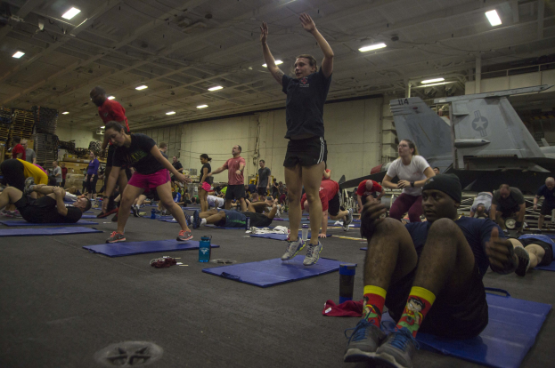 Eine Gruppe von Menschen, die Yoga in einem großen Raum mit einer Wand, Deckenlampen und verstreuten Gegenständen, einschließlich Schuhen, praktizieren.