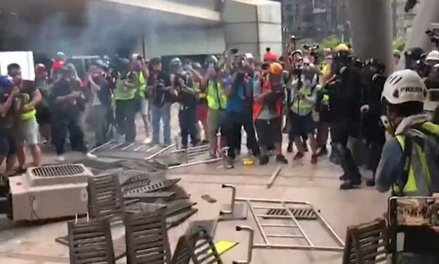 Menschen vor einem rauchenden Gebäude in einer städtischen Umgebung, einige mit Helmen und Jacken, andere mit Kameras, mit verstreuten Metallobjekten und Trümmern auf dem Boden.