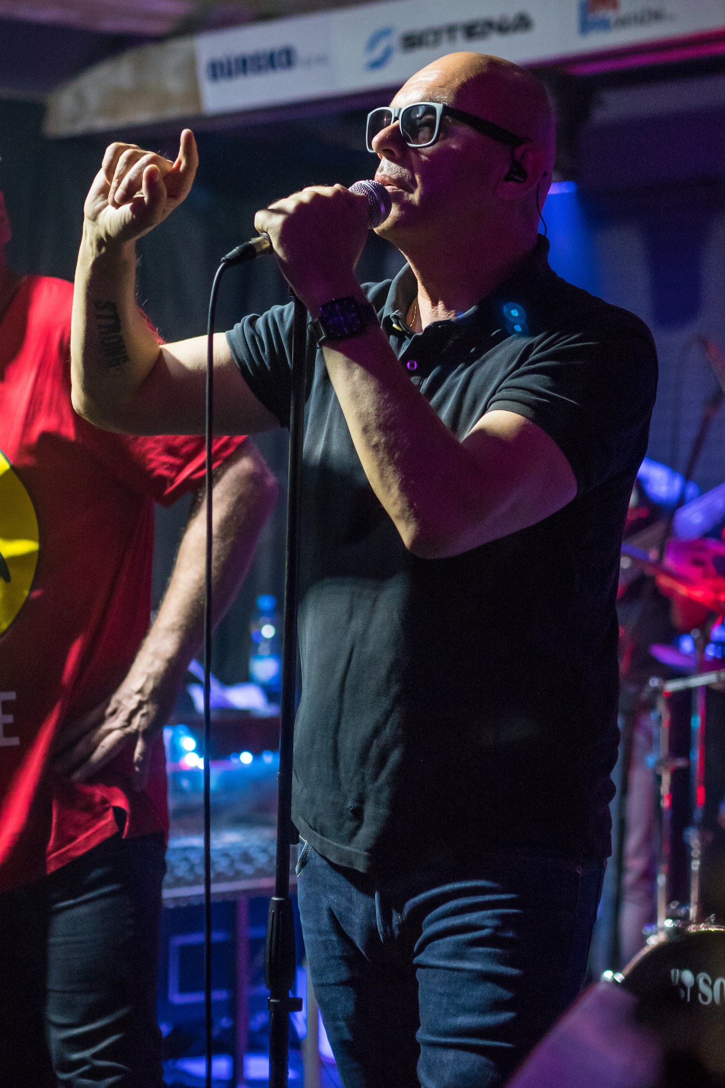 Ein Mann singt in ein Mikrofon auf der Bühne einer Veranstaltung, trägt ein schwarzes T-Shirt und blaue Jeans und hat einen anderen Musiker mit einem Instrument hinter sich.