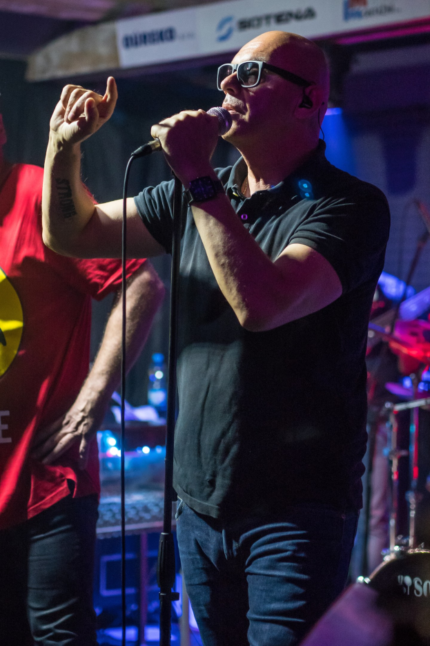 Ein Mann singt in ein Mikrofon auf der Bühne einer Veranstaltung, trägt ein schwarzes T-Shirt und blaue Jeans und hat einen anderen Musiker mit einem Instrument hinter sich.