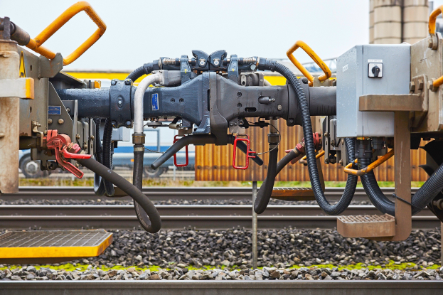 Nahaufnahme einer Bahnstrecke mit einer Bohrmaschine, umgeben von Schotter und Gras, mit einem Fahrzeug, einem Gebäude und dem Himmel im Hintergrund.