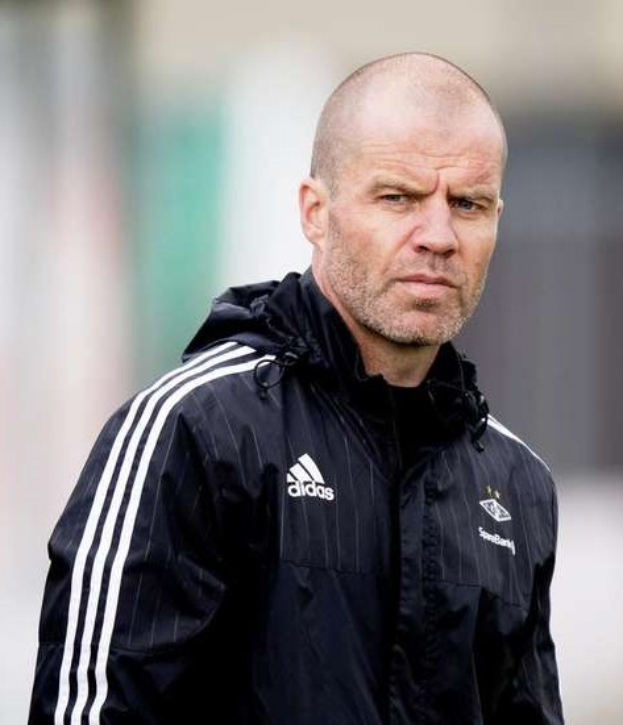 Mann mit Glatze in einem blauen Jackett mit weißen Streifen und einem weißen Kragen, der auf einem Fußballfeld steht und die Hände verschränkt hält.