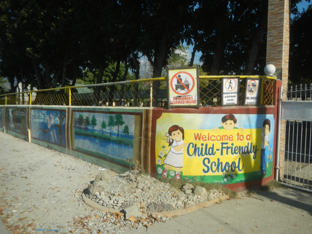 Eine Wand mit einem "Willkommen in einer kinderfreundlichen Schule"-Schild, umgeben von einem Metallzaun, einer Säule, Bäumen, Steinen und einem Weg.