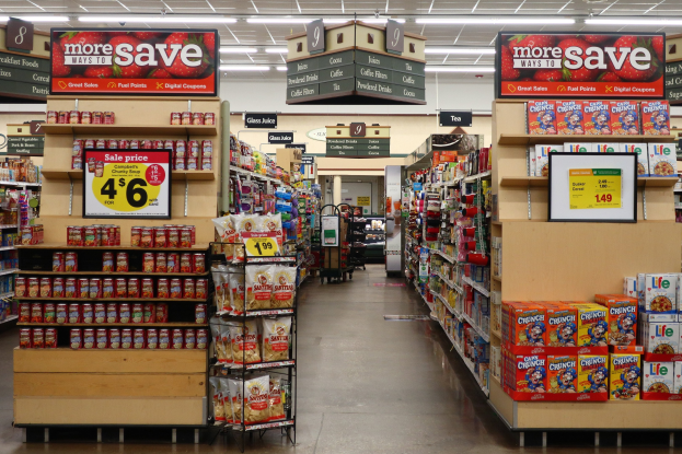 Ein Kiosk-Innenraum mit Regalen und Boden voller verschiedener Lebensmittel, Deckenbeleuchtung und Informationsschilder im Hintergrund mit einer Person im Hintergrund.