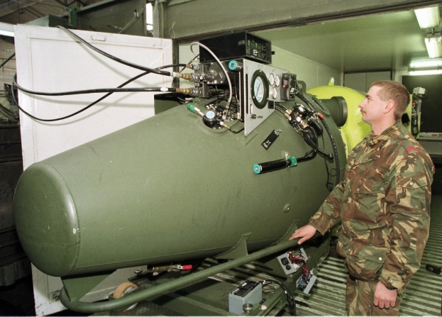 Ein Mann in militärischer Uniform steht neben einer großen grünen Bombe in einer unordentlichen industriellen Umgebung mit Maschinen, Kabeln und beleuchtetem Hintergrund.