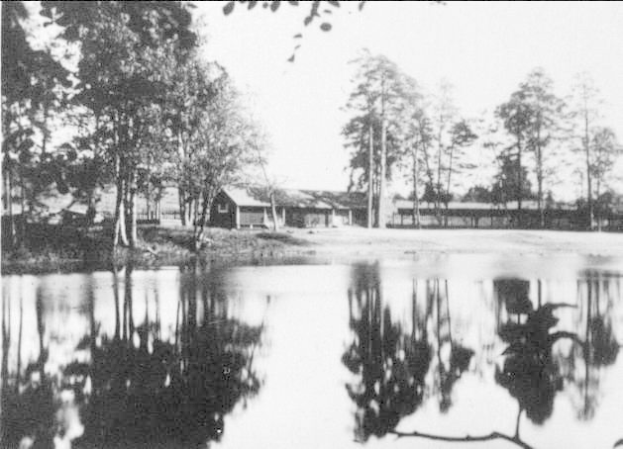 Schwarzes und weißes Foto eines Sees mit Bäumen im Hintergrund, umgeben von Häusern und einem klaren Himmel, mit Text unten, der "Lakeside Lodge in den 1930er Jahren" lautet.