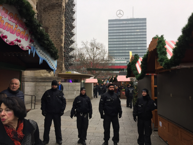 Eine Gruppe von Polizisten in schwarzen Uniformen und Mützen steht vor einem geschmütckten Weihnachtsmarkt in Berlin, mit festlichen Dekoelementen, Gebäuden, Bäumen und einem klaren blauen Himmel im Hintergrund und einer Tafel mit Text links.