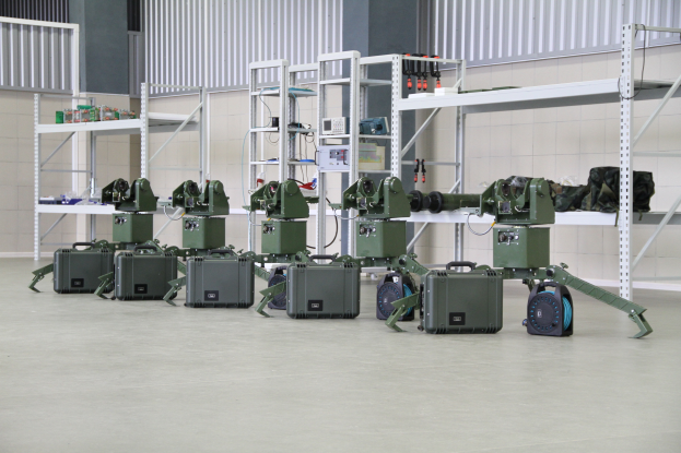 Eine Gruppe von Militärrobotern auf einem Boden umgeben von Koffern und anderen Gegenständen, mit einer Wand und einem Geländer im Hintergrund.